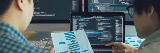 Two engineers examining a process flow on paper with coding on computer screens in the background.