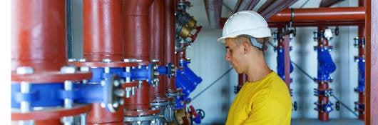 A male heating plant worker wearing a hardhat is standing next to a pipe system and performing a quality check.