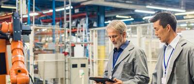 Two engineers walking around in a factory with a digital tablet and talking. Robotic arm in the background.